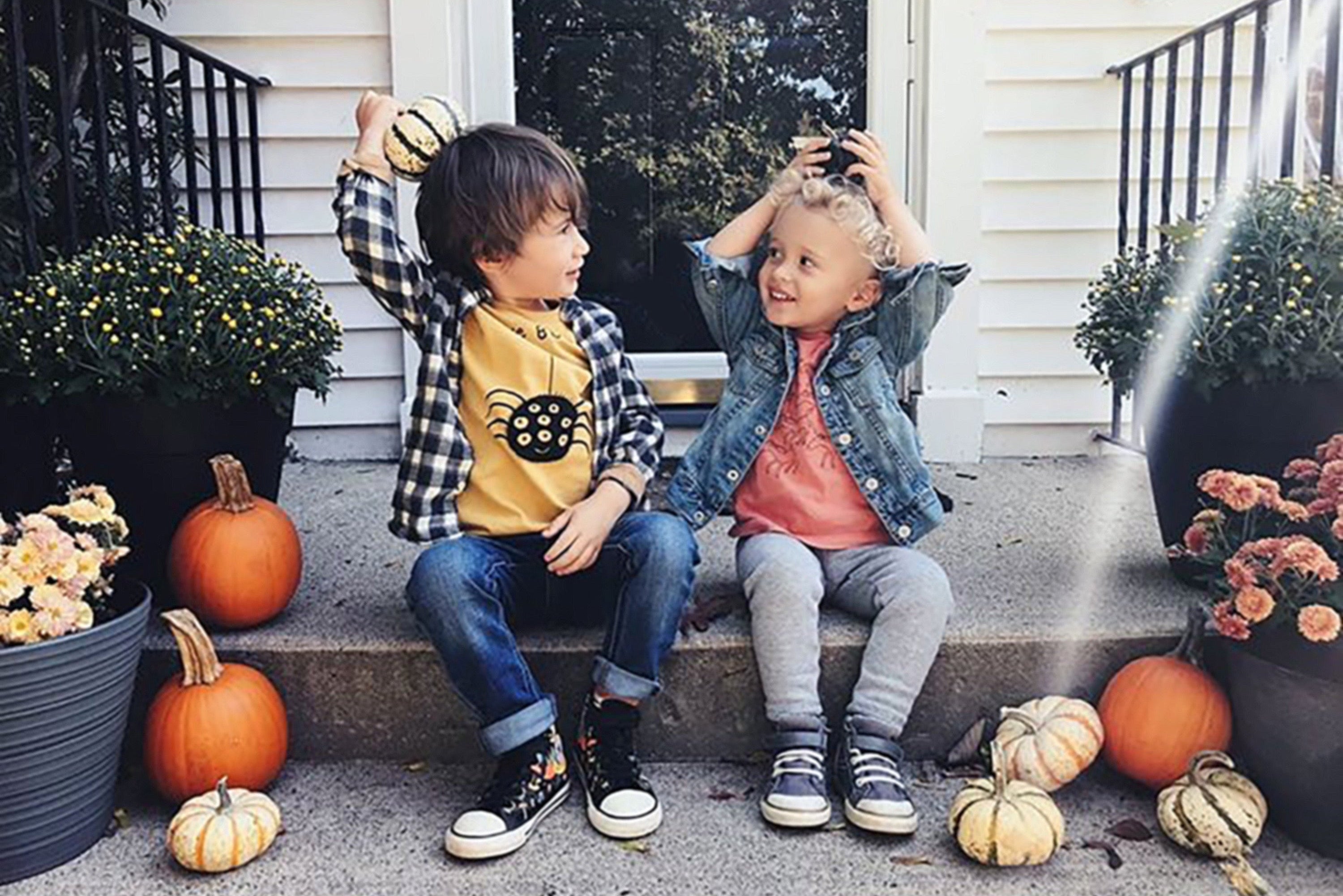 Two children sitting on a porch with pumpkins and flowers around them.