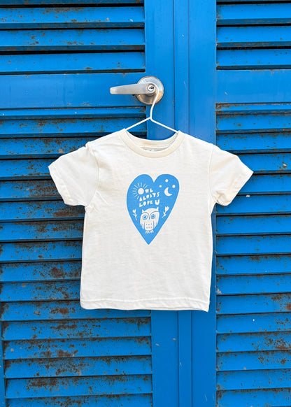 Natural organic cotton t-shirt with a heart and owl on it, hanging on a door handle of a blue rusty door.